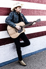 Old cowboy with gray beard plays a guitar while standing next to a white and red wall.