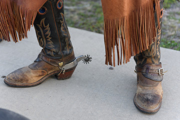 Old cowboy boots are weathered and brown with stitched design on the side with spurs worn by an old cowboy in jeans.