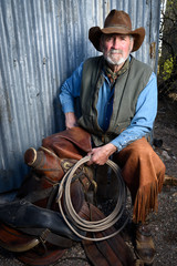 Old cowboy with gray beard wears leather hat, leather chaps, and holds a lasso and is sitting with a leather saddle.