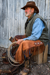 Old cowboy with gray beard wears leather hat, leather chaps, and holds a lasso and is sitting with...