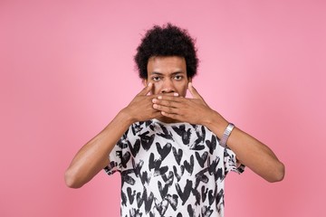 Curly African man covering his mouth with his hands without saying a word standing on a pink background