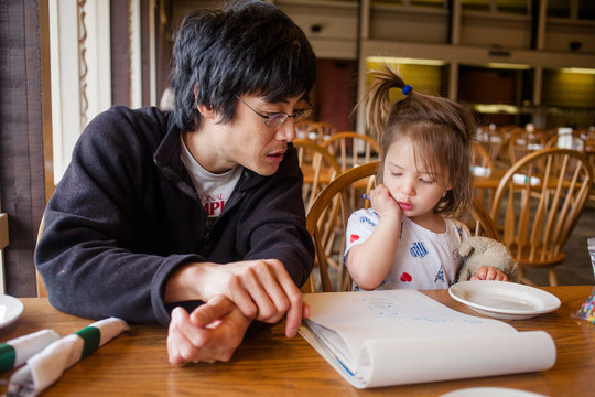 A father and little girl draw pictures at a restaurant table by window