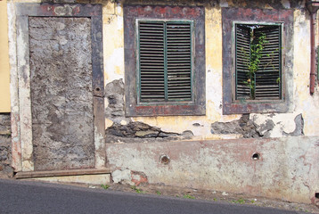 an old ruined house partly collapsed on a sloping street with a blocked up door crumbling walls and fading red painted windows with green closed shutters