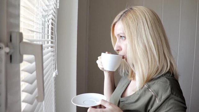 Young sad thinkful beautiful blond woman standing near the window with blinds in the morning and drinking coffee