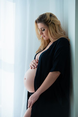 A beautiful pregnant woman in a black robe stands near a bright window. Feminine sexuality, femininity and beautiful pregnancy