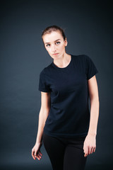 Obraz premium sporty-looking girl in a black T-shirt and black leggings on a dark background and slightly bent looking at the camera