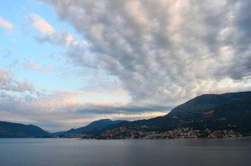 A bay seen at sunset. Grey, pink and white clouds stretch across a blue sky. Rocky hills with mountains in the background surround the bay.