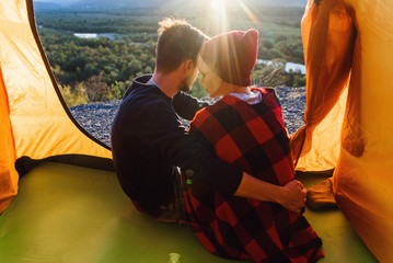 View from the tent of young caucasian couple in love while they sitting on the mountains background at sunset. View fro back.