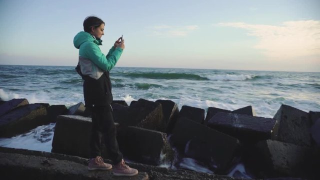 attractive woman in a warm jacket making photo on smart phone at sunset near sea. Slow motion