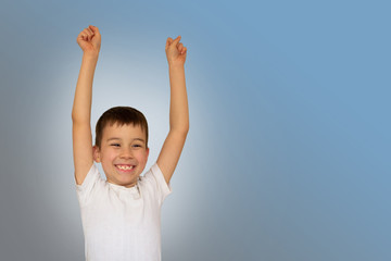 A boy in a light T-shirt celebrating a happy, smiling laugh with raising hands on a blue background, copy space, close-up