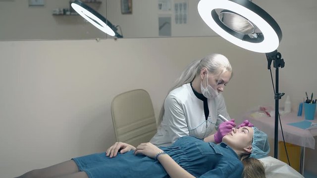 girl in blue dress lies on couch and looks at cosmetician making permanent make-up in beauty salon