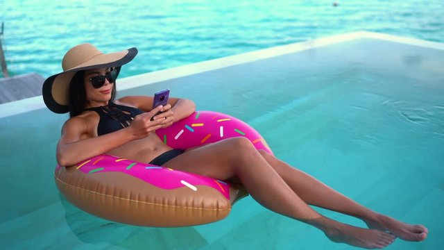 Summer Vacation Woman in bikini on inflatable donut mattress using mobile cell phone in swimming pool. Top view of girl relaxing sunbathing enjoying travel holidays at resort pool. Luxury lifestyle.