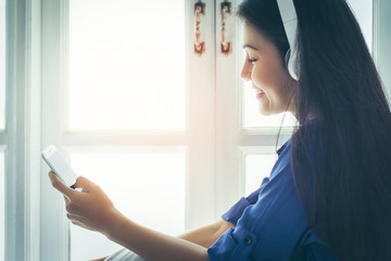 Asian pretty girl wearing blue shirt sitting by the window in the bedroom is white. She smiled happily sat listening to music from headphones which audio connector from the smartphone with copy space.