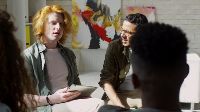 Waist-up shot of Caucasian guy with bright red hair and Middle Eastern man wearing glasses tentatively talking to colleagues at informal work meeting in sunny vibrant creative design studio