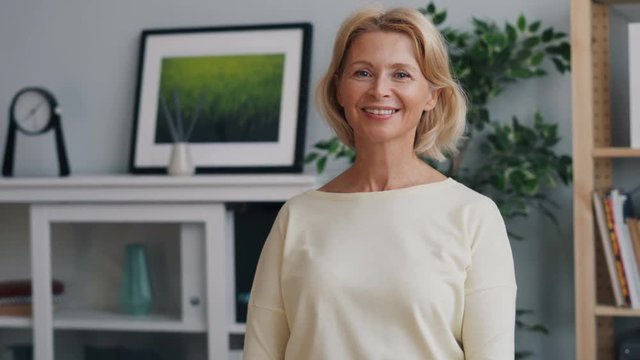 Portrait of happy mature lady looking at camera and smiling standing in office alone with shelves and pictures in background. Cheerful people and business concept.