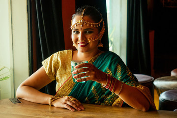 pretty Indian/Asian girl in sari holding glass of plain water, sitting at home kitchen near window