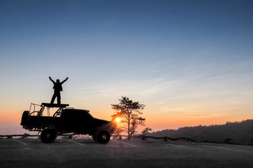 Hug the sky, view silhouette of a tourist standing on roof top of a 4WD. truck with colorful vivid sun light in the sky background, sunset at Huai Nam Dang National Park, Chiang Mai, Thailand.