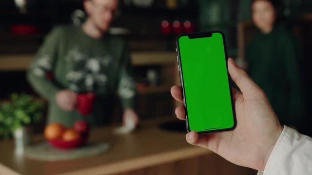 Young hands hold use smartphone with vertical green screen on kitchen at home background people quarrel breakfast apple fruit browse house shopping internet finger payment communication close up