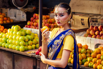 indian Caucasian woman in traditional dress.hindu model golden kundan jewelry set bindi earrings and nose ring piercing nath choosing fruits shopper in a supermarket fruit shop.exotic sale Kerala Goa