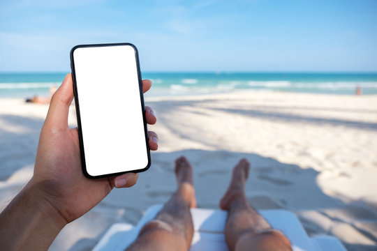 Mockup image of a man's hand holding white mobile phone with blank desktop screen while laying down on beach chair