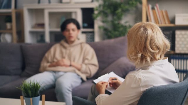 Blond woman professional psychologist is consulting unhappy teenager in clinic. Boy is sitting on couch talking and gesturing while doctor is giving advice.