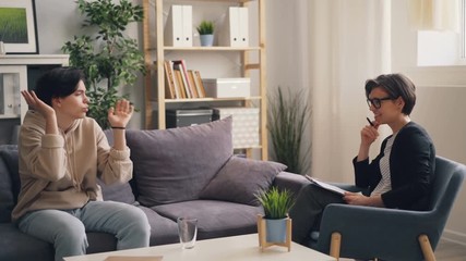 Emotional teen with dark hair is discussing personal troubles with young woman therapist sitting on couch in office. Lady is listening and giving advice.