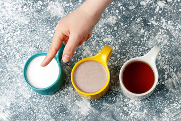 Multicolored mugs with hot drinks and female hand on a gray background, top view