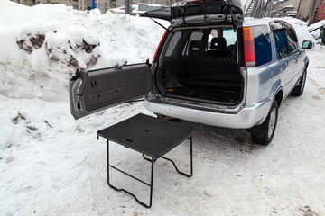 SUV car in silver color with opened trunk and table for picnic near it after cleaning before sale in a winter day and snow background