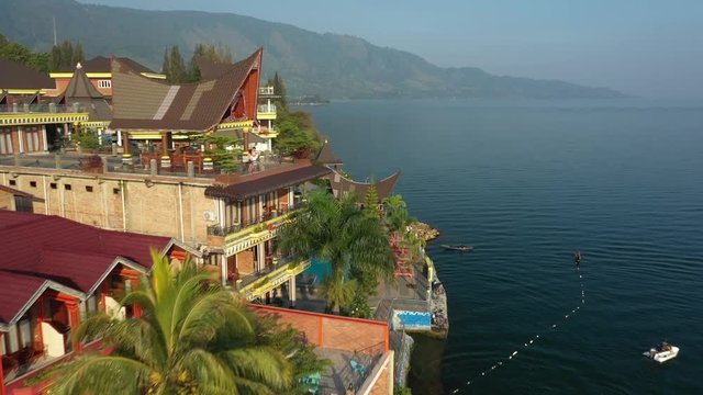 Flyover drone footage of lakeside resorts with traditional Batak roofs on Lake Toba in North Sumatra, Indonesia.