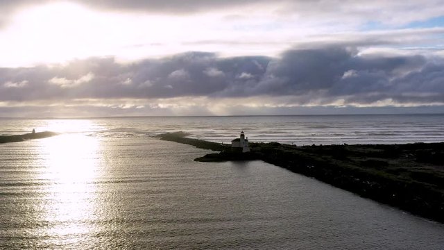 Beautiful minimalistic landscape image of drone flying over the Coquille river in Bandon Oregon towards the lighthouse, which is silhouetted from the sunset behind it.