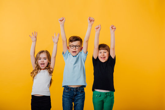 three beautiful children, little boy in glasses and two little girls jumping up, hands raised, happy, glad, isolated yellow background, copy space