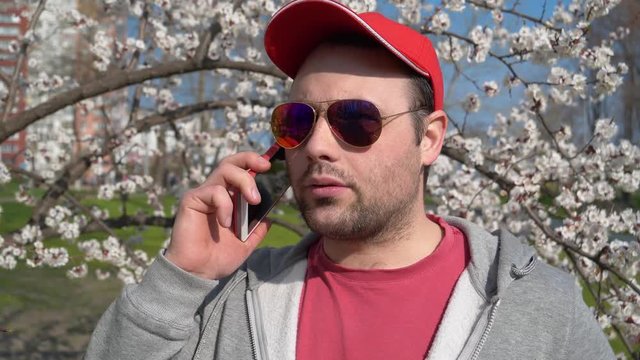Man in red baseball cap with red smartphone. Emotional man in sunglasses talks on smartphone in city park