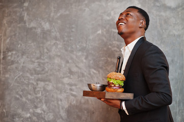 Respectable young african american man in black suit hold tray with double burger against gray wall.