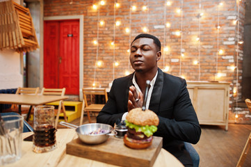 Pray before eat. Respectable young african american man in black suit sitting in restaurant with tasty double burger and soda drink.