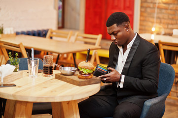 Looking at wallet to find cash for pay. Respectable young african american man in black suit sitting in restaurant with tasty double burger and soda drink.