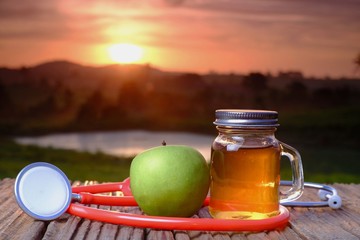 Stethoscope, apple vinegar and green apple on wooden table with beautiful sunrise as background