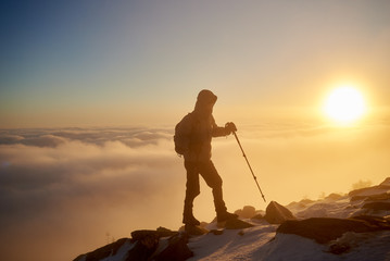 Silhouette of tourist hiker with backpack and hiking sticks walking on rocky mountain top on copy space background of beautiful foggy valley filled with white puffy clouds at sunrise.