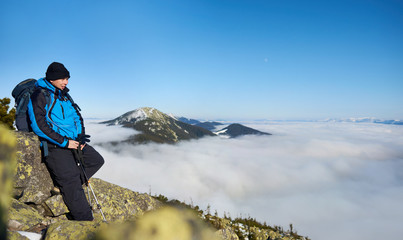 Tourist hiker with backpack and trekking poles resting on steep rocky mountain slope on background of blue sky, foggy valley filled with white clouds and snowy mountain tops.