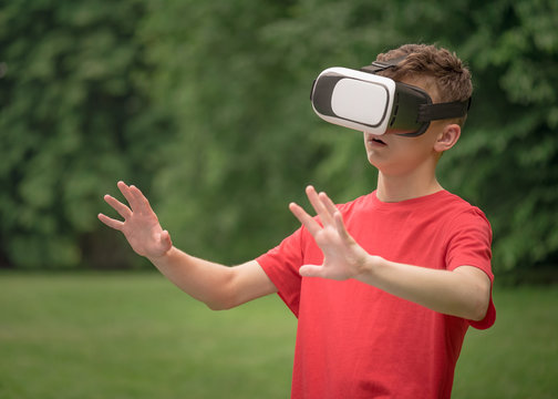 Playful surprised teen boy using virtual reality goggles outdoor in summer park. Teenager looking in VR glasses. Child have fun - experiencing 3D gadget technology.