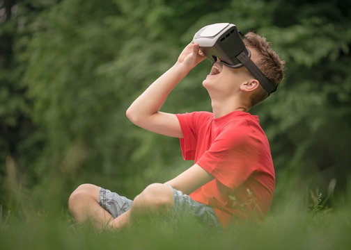 Playful teen boy using virtual reality goggles outdoor in summer park. Teenager looking in VR glasses. Child have fun - experiencing 3D gadget technology.