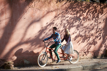 Young happy tourist couple in casual clothing, bearded man and blond woman in glasses cycling tandem bike along empty paved sunny sidewalk on background of high red crackled plastered wall.