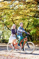 Fototapeta premium Young tourist pair, handsome bearded man and attractive blond long-haired woman cycling together tandem double bike by sunny alley with golden leaves on tall trees background in the fall