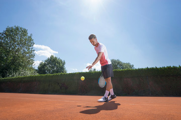 Young male tennis player preparing to serve the ball on sunny red tennis court, Bavaria, Germany