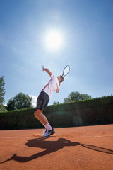Young male tennis player serving the ball on sunny red tennis court, Bavaria, Germany