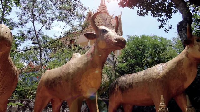 Golden magical animals including a bull, rooster, and parrot guard a Buddhist temple in the forest of Southeast Asia. Camera is low aerial shot (drone), and flies right to reveal more animals and stup