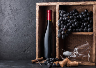 Bottle of red wine and empty glasses with dark grapes inside vintage wooden box on black stone...