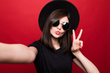 Young woman in sunglasses, black hat and black clothes take selfie on phone on red background