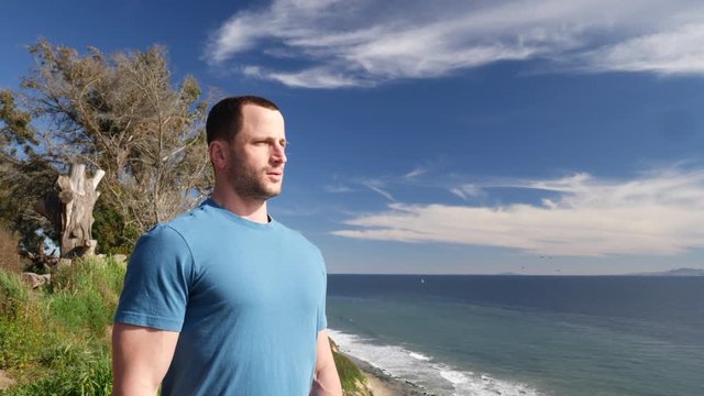 A man taking a deep breath and meditating to relive stress on an epic ocean cliff above the beach waves in Santa Barbara, California SLOW MOTION.