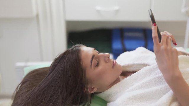 Young Caucasian Female Laying Down on Bed Between Hair Mesotherapy Treatments and Looking At Smartphone. Light Camera Movement
