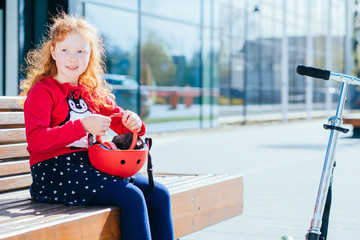 Portrait of cute adorable smiling little curly red-haired caucasian girl child sitting on bench,...